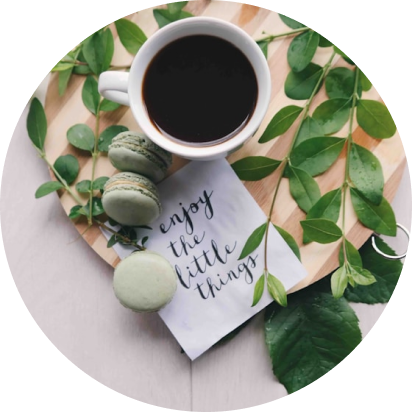 A cup of coffee, macarons, and a note that says 'enjoy the little things' on a table with green leaves